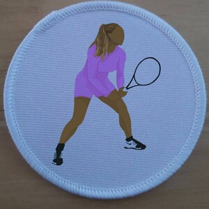 May include: A white round patch with a pink tennis player holding a black tennis racket. The player is wearing a pink outfit and black shoes.