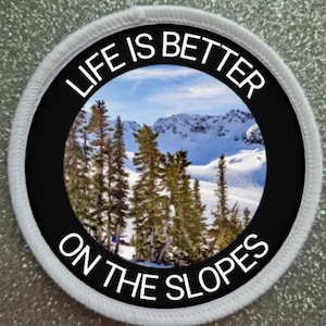 Op de afbeelding: Een ronde witte patch met zwarte tekst die "Life is better on the slopes" zegt en een foto van een besneeuwde berg scène met bomen.