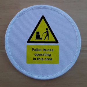 May include: A white round warning sign with a yellow triangle and a black stick figure pushing a pallet jack. The text below the triangle reads "Pallet trucks operating in this area."
