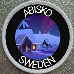 Op de afbeelding: Een witte ronde patch met zwarte tekst die "Abisko Zweden" luidt. Het midden van de patch toont een zwarte cirkel met een kleurrijk beeld van een besneeuwd huis met een geel raam en een paarse aurora borealis in de lucht.