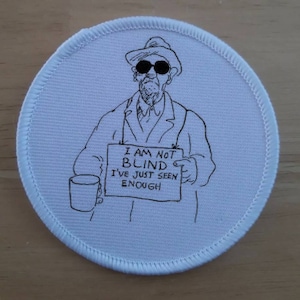 May include: A white round patch with a black and white illustration of a person wearing a hat and sunglasses holding a sign that reads "I am not blind I've just seen enough".