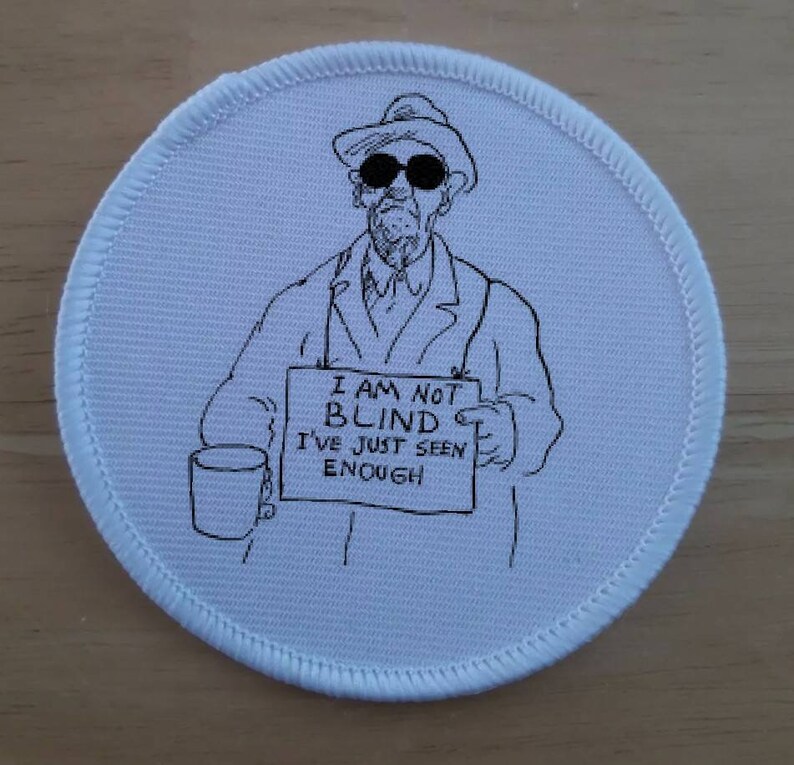 Puede incluir: Un parche redondo blanco con una ilustraci&oacute;n en blanco y negro de una persona con sombrero y gafas de sol sosteniendo un cartel que dice "No estoy ciego, solo he visto suficiente".