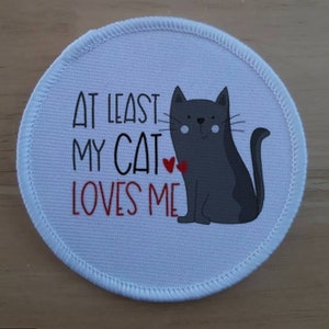 May include: White round fabric patch with a grey cat illustration and the text "At least my cat loves me".