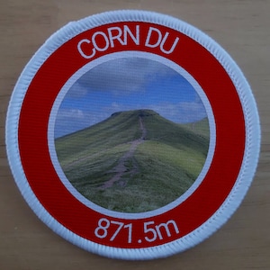 May include: A white and red circular patch with the text "CORN DU" and "871.5m" in white. The centre of the patch features a photo of a green mountain.