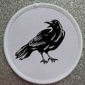 Può includere: Una toppa ricamata in bianco e nero di un corvo con il becco aperto, su uno sfondo circolare bianco.
