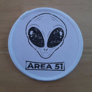Puede incluir: Parche redondo blanco con una cara de extraterrestre en blanco y negro y el texto "AREA 51" impreso en él.