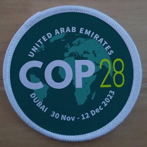 Op de afbeelding: Een ronde groene en witte patch met de tekst "UNITED ARAB EMIRATES COP28 DUBAI 30 Nov - 12 Dec 2023" en een groene omtrek van de wereld.