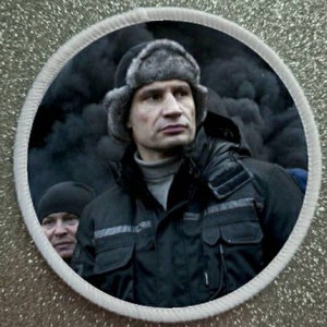 Può includere: Un uomo con un cappello di pelliccia grigio scuro e una giacca nera con una cerniera grigia. È in piedi davanti a uno sfondo scuro.