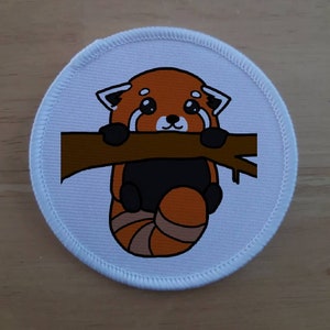 Könnte beinhalten: Ein weißer runder Patch mit einem Roten Panda, der sich an einem braunen Ast festhält. Der Rote Panda hat ein weißes Gesicht mit schwarzen Augen und einer schwarzen Nase. Das Fell des Roten Pandas ist orange und braun.