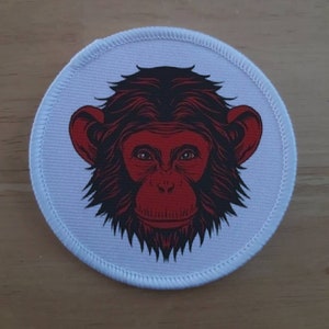 Könnte beinhalten: Ein weißer runder Patch mit einer schwarzen und roten Illustration eines Affengesichts.
