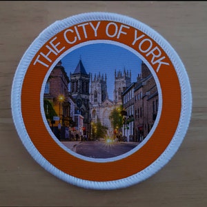 Puede incluir: Un parche redondo de color naranja y blanco con el texto "THE CITY OF YORK" en letras blancas alrededor de una foto de una calle en York, Inglaterra. La foto muestra una calle con edificios a ambos lados y una iglesia en el fondo.