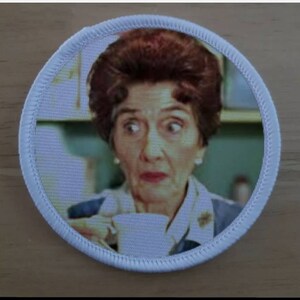May include: A circular embroidered patch featuring a photographic print of a person with dark brown hair styled in a bouffant, wearing a collared shirt, and holding a white teacup. The patch has a thick white stitched border around the edge.