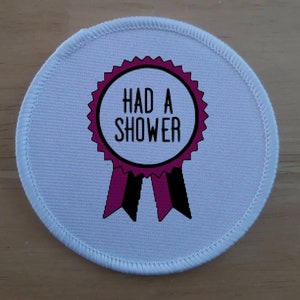 Könnte beinhalten: Runder Stoff-Patch in Weiß mit einem rosa und schwarzen Band-Award-Design. Das Band trägt den Text "HAD A SHOWER" in Schwarz.