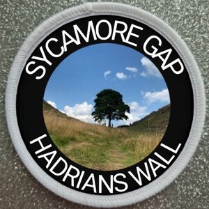 Op de afbeelding: Een witte ronde patch met zwarte tekst en een zwarte cirkel in het midden. De tekst luidt "SYCAMORE GAP" en "HADRIANS WALL". De zwarte cirkel bevat een foto van een boom in een veld met een blauwe lucht en witte wolken.
