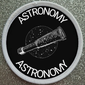Może przedstawiać: Okrągła łatka w kolorze białym i czarnym z napisem "ASTRONOMIA" napisanym dwa razy wokół krawędzi. W środku łatki znajduje się czarno-biała ilustracja teleskopu.