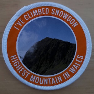 Może przedstawiać: Okrągła naszywka w kolorze białym i pomarańczowym z napisem "I've Climbed Snowdon Highest Mountain in Wales". Naszywka zawiera zdjęcie szczytu góry.