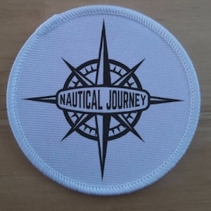 以下が含まれることがあります： 黒いコンパスデザインと中央に「NAUTICAL JOURNEY」というテキストが書かれた白い丸い布のパッチ