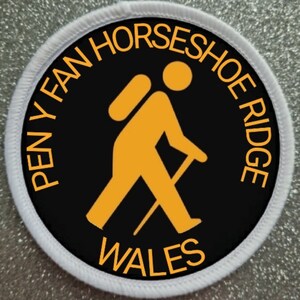 May include: A black and white round patch with orange text and a silhouette of a hiker with a backpack and walking stick. The text reads "Pen y Fan Horseshoe Ridge Wales".