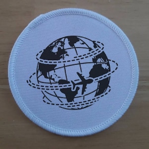 May include: A circular white patch featuring a black graphic of the Earth, encircled by dotted lines, and a plane silhouette. The patch has a white stitched border, possibly an emblem or decorative item.