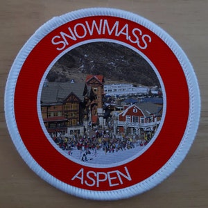 Puede incluir: Un parche circular blanco y rojo con las palabras "SNOWMASS ASPEN" en texto blanco. El centro del parche presenta una foto de un pueblo de montaña nevado.