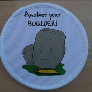 Può includere: Toppa rotonda bianca con un'immagine di cartone animato di due massi e una piccola tenda. Il testo "Another year boulder!" è stampato in nero nella parte superiore della toppa.