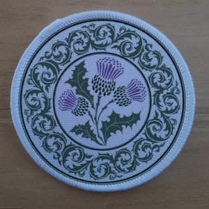 Pode incluir: Um remendo redondo de tecido branco com um design de cardo verde e roxo. O cardo é rodeado por uma borda verde decorativa.