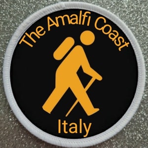 Può includere: Un cerotto rotondo nero e bianco con un bordo bianco. Il cerotto presenta una figura stilizzata gialla di un escursionista con uno zaino e un bastone da trekking. Il testo "The Amalfi Coast Italy" è scritto in giallo attorno all'escursionista.