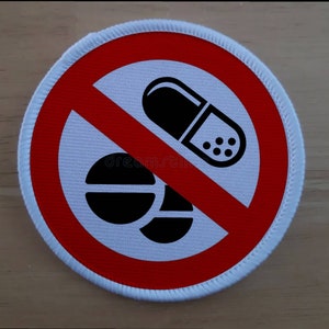 Puede incluir: Un parche circular blanco y rojo con una línea diagonal roja en el centro. El parche tiene una imagen negra de una píldora y dos tabletas. El texto "No Drugs" no es visible en la imagen.