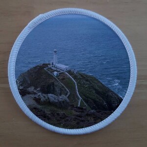 May include: A white lighthouse sits on a rocky cliff overlooking a blue ocean. The lighthouse is surrounded by green grass and a winding path.