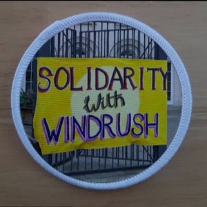 Puede incluir: Un parche circular blanco con una pancarta amarilla que dice "Solidaridad con Windrush". La pancarta está colgada de una puerta de metal negro.