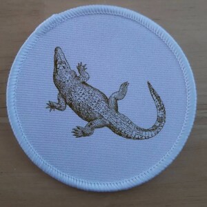 May include: White round fabric patch with a brown alligator illustration.