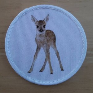 May include: A white fabric patch with a brown and white fawn printed on it. The fawn is standing on all four legs and looking directly at the camera.