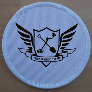 May include: White round patch with black embroidered design. A shield with wings and a metal detector and shovel crossed inside. The text "TREASURE HUNTER" is on a banner below the shield.