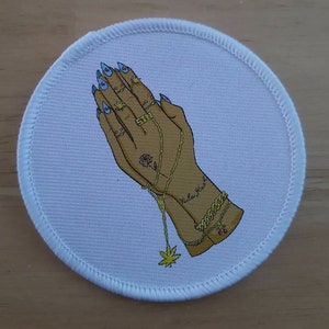 May include: A white round patch with a colorful illustration of two hands clasped in prayer. The hands are adorned with gold rings, bracelets, and a necklace with a marijuana leaf pendant. The text "Vuela High" is written on one of the wrists.