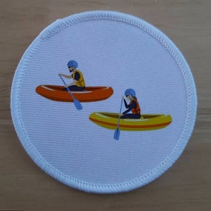 May include: A white round patch with two people in canoes. The person in the orange canoe is wearing a blue helmet and a yellow life jacket. The person in the yellow canoe is wearing a blue helmet and a red life jacket.
