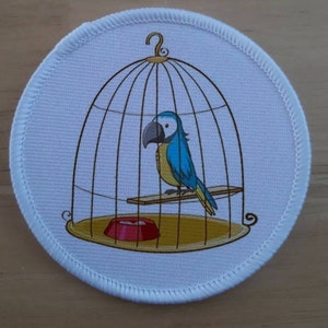 May include: A blue and yellow parrot in a cage with a red food bowl. The cage is made of metal and has a wooden perch.