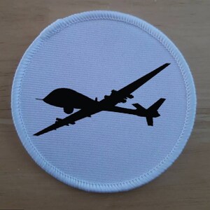 Op de afbeelding: Ronde patch met een zwart silhouet van een drone op een lichtblauwe achtergrond. De patch heeft een witte gestikte rand. De drone heeft een lange, slanke romp en vleugels.
