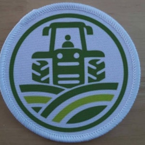 May include: A white round patch with a green outline and a green tractor in the centre. The tractor is driving over a green field with yellow stripes.