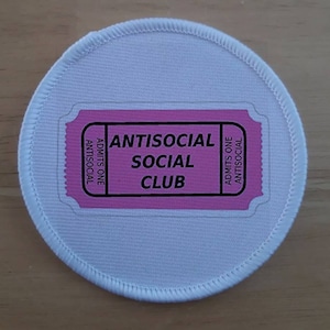 May include: White round patch with a pink ticket that says "Antisocial Social Club" in black text.