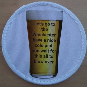 Può includere: Cerotto rotondo bianco con un bicchiere di birra. Il testo sul cerotto recita: "Let's go to the Winchester, have a nice cold pint, and wait for this all to blow over."