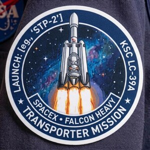 May include: A circular embroidered patch featuring a SpaceX Falcon Heavy rocket launching into a blue and purple nebula. The text reads LAUNCH: leg., 'STP-2', KSC LC-39A, SPACEX, FALCON HEAVY, and TRANSPORTER MISSION with white stars on a dark blue border.