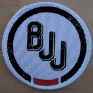 Puede incluir: Parche redondo blanco con un círculo negro y el texto "BJJ" en letras negras.