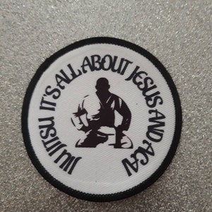 May include: White round patch with black trim. The patch features a black silhouette of a person in a martial arts pose. The text around the person reads "It's all about Jesus and Jiu Jitsu".