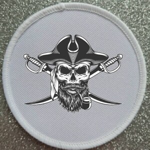 Könnte beinhalten: Ein weißer runder Patch mit einer Schwarzweißillustration eines Schädels, der einen Piratenhut trägt und eine Pfeife hält. Zwei gekreuzte Schwerter befinden sich hinter dem Schädel.