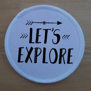 May include: White round fabric patch with black stitching around the edge. The patch has a black arrow pointing to the right and the text "Let's Explore" in a stylized font.
