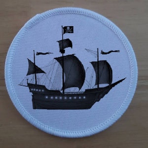May include: A white round patch with a black and white image of a pirate ship with a skull and crossbones flag. The ship has three masts and sails.