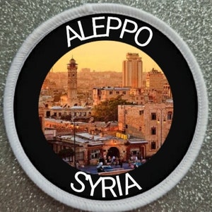 May include: A round, white patch with a black circle in the centre. The circle features a colourful image of the city of Aleppo, Syria, with the words "Aleppo" and "Syria" printed in white at the top and bottom of the circle.