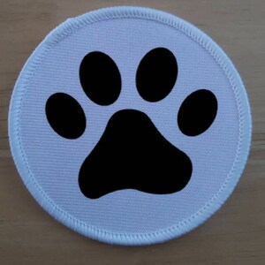 Könnte beinhalten: Ein weißer Stoff-Patch mit einem schwarzen Pfotenabdruck. Der Pfotenabdruck befindet sich mittig auf dem Patch.
