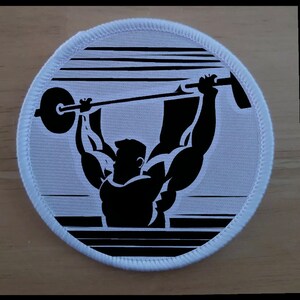 May include: White circular patch with a black silhouette of a weightlifter lifting a barbell. The weightlifter is in a classic weightlifting pose with a striped background.