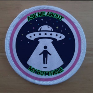 May include: A white round patch with a pink border. The patch has a dark blue background with a white UFO and a white stick figure standing in the beam of light from the UFO. The text "ASK ME ABOUT MY BUTTHOLE" is written in green around the edge of the patch.